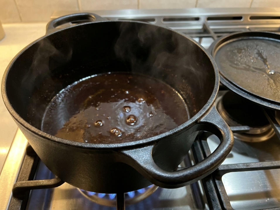Black enameled cast iron dutch oven on a stovetop burner with sauce reducing inside and steam rising, lid resting beside it